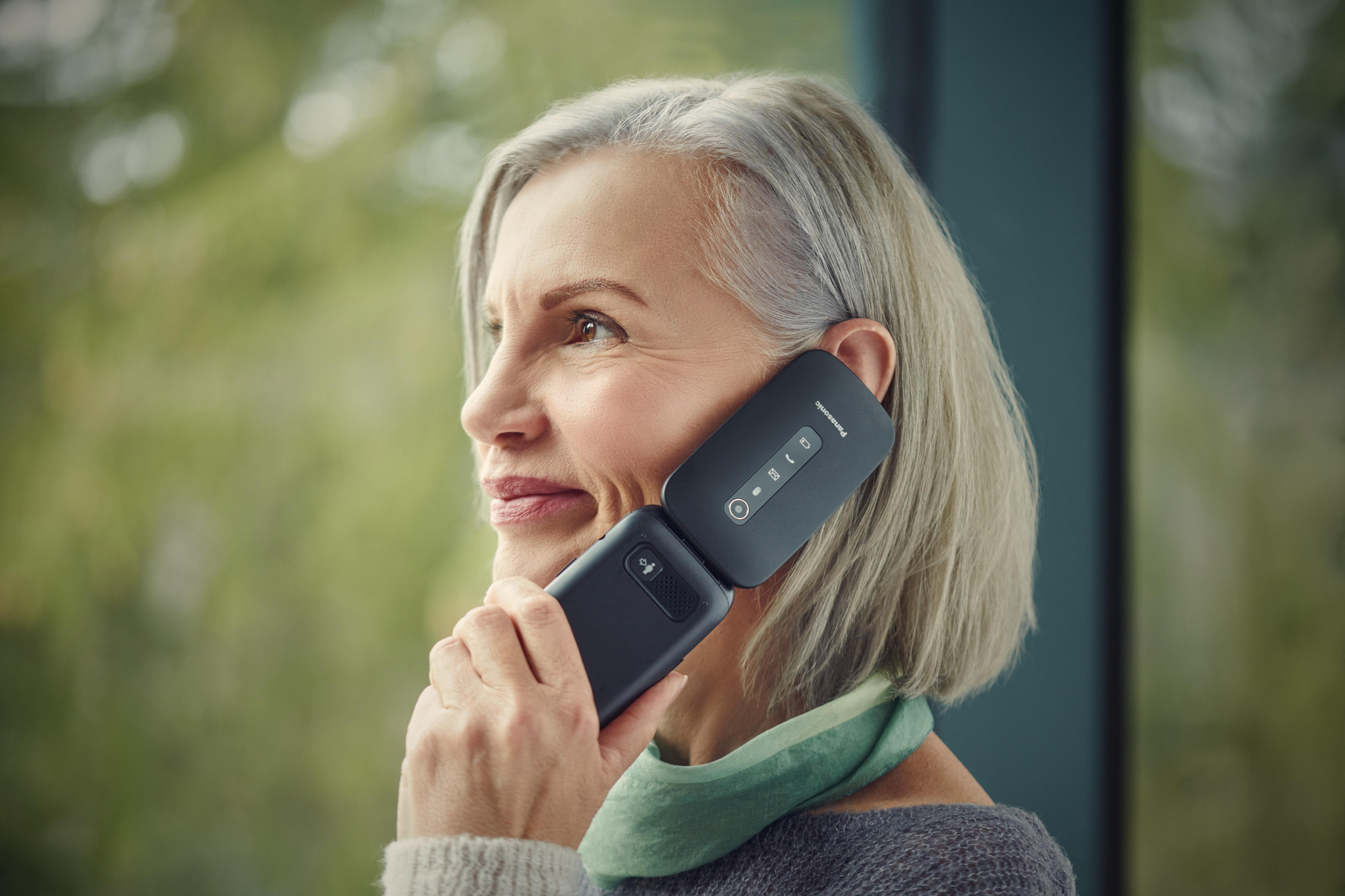 Eine Frau mit grauem Haar hält ein schwarzes Telefon an ihr Ohr und lächelt. Grüner Schal um den Hals. Verschwommener Hintergrund.