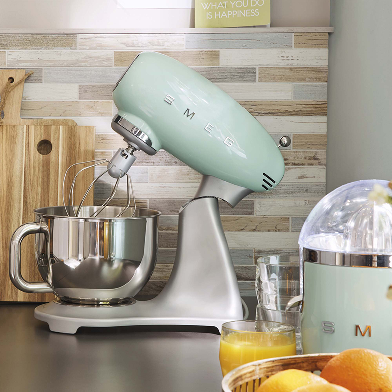 Un mixer a immersione Smeg verde menta con una ciotola in acciaio inox, accanto a uno spremiagrumi e taglieri in legno.