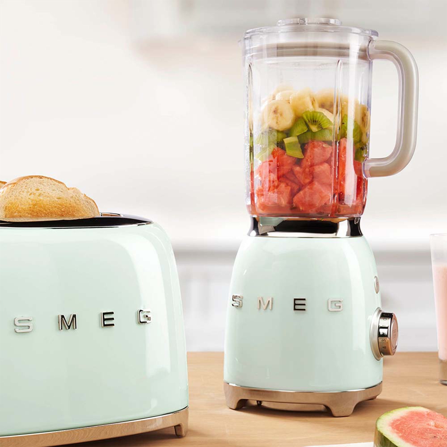 Un tostapane e un frullatore SMEG verde menta con frutta all'interno, vicino a un tostapane con pane.