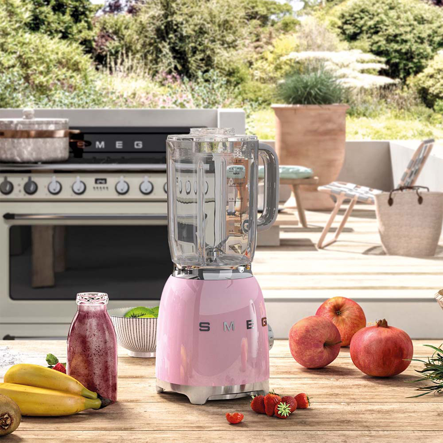 Frullatore rosa con frutta e verdura su un tavolo di legno all'aperto.