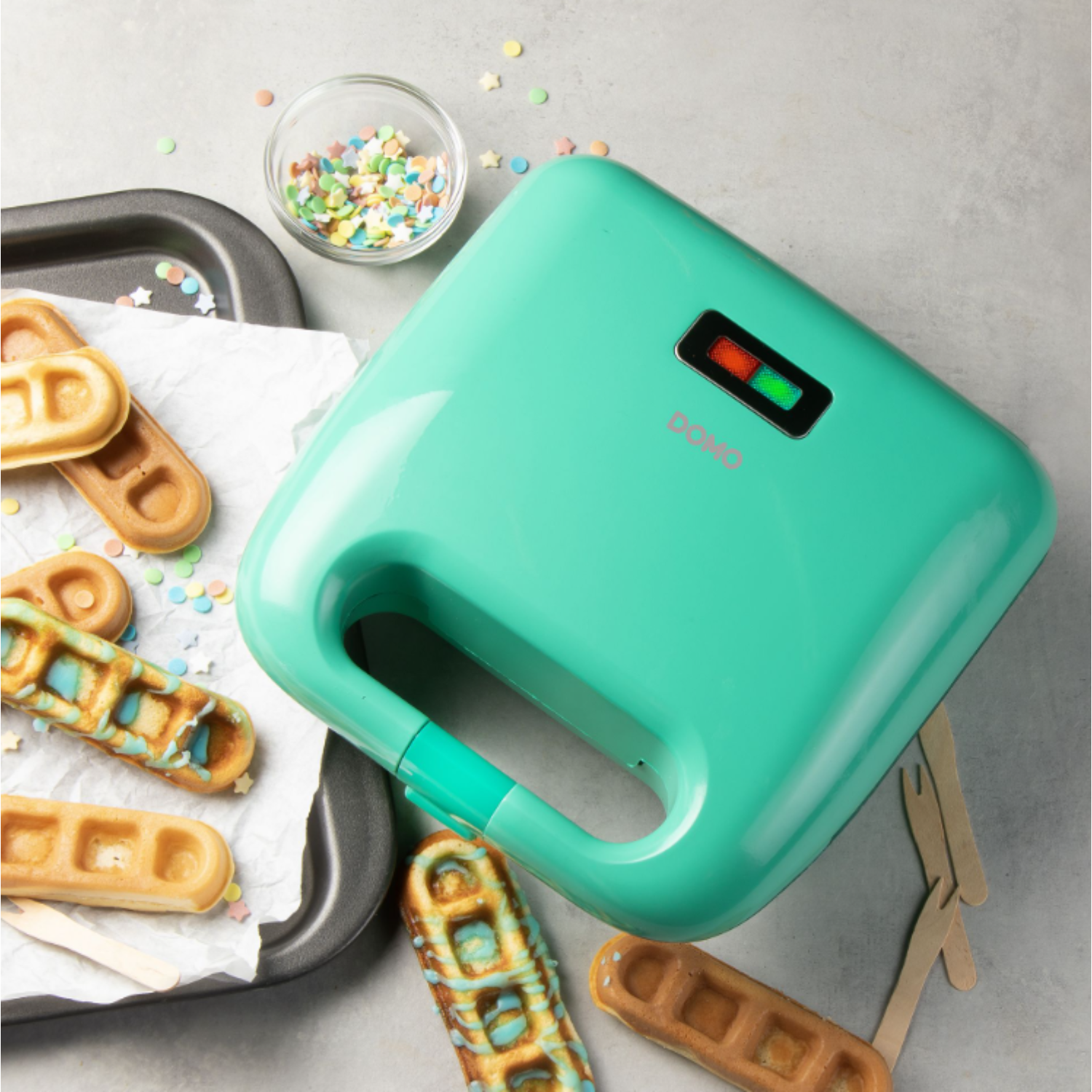 A teal waffle maker with waffles, sprinkles, and a tray. Green light on.