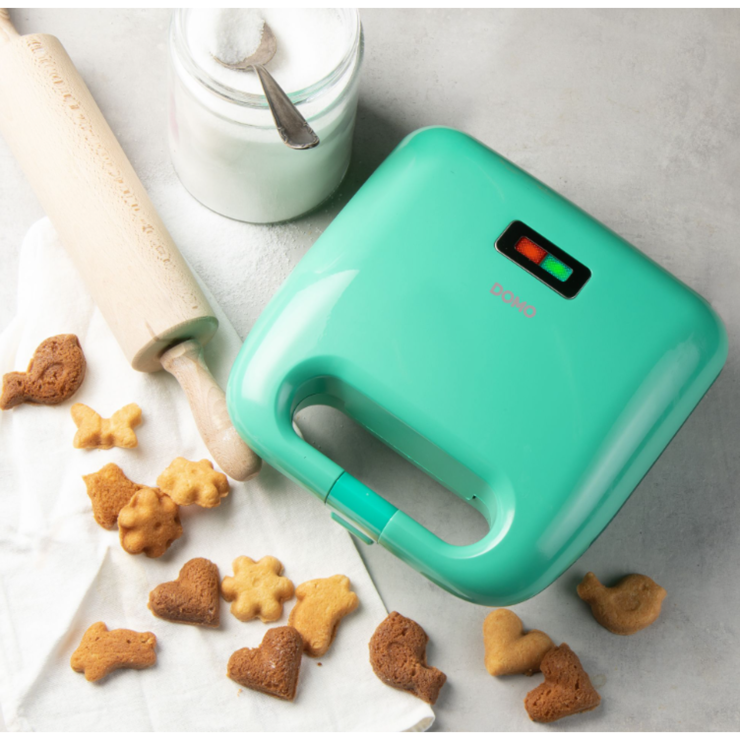A green waffle maker with cookies, rolling pin, and sugar jar on a table.