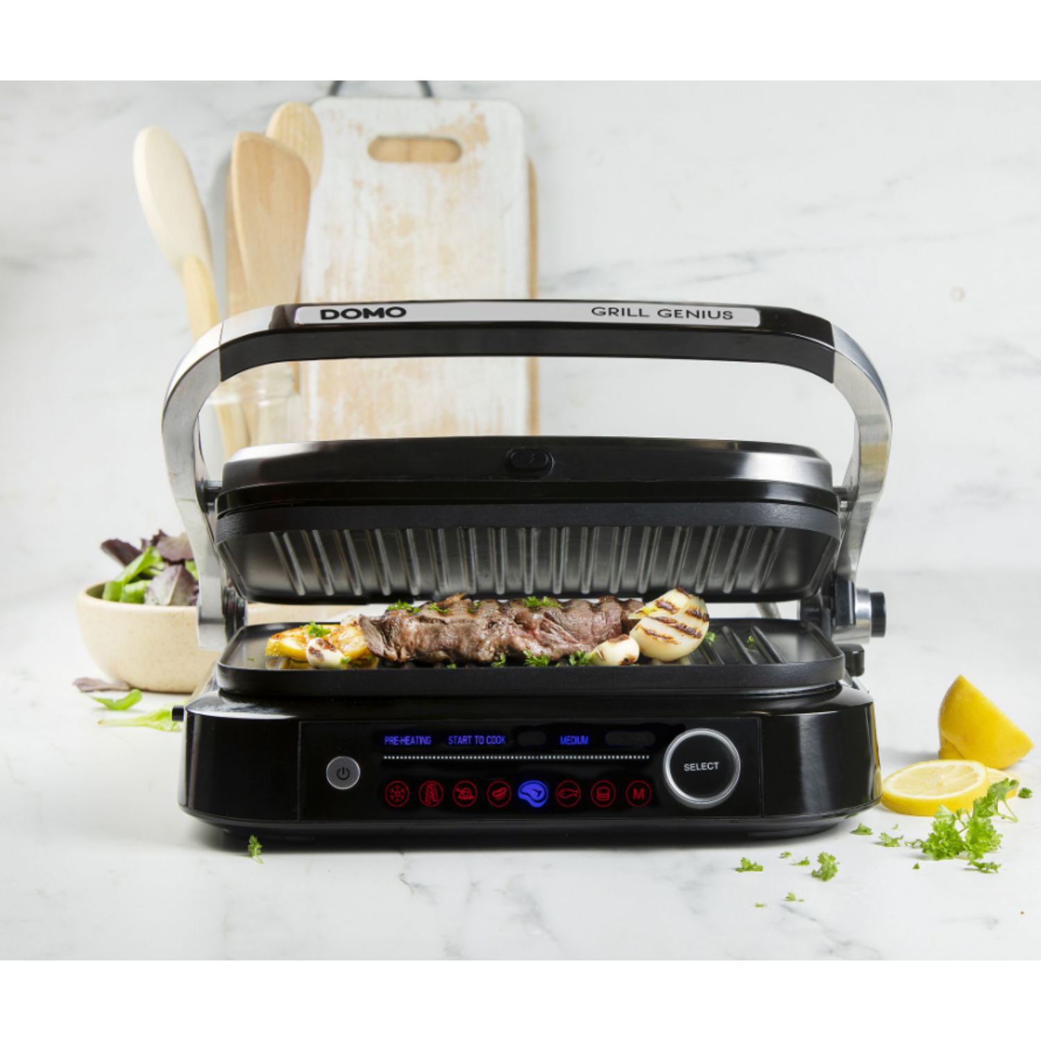 A grill with food cooking on it. There are lemons, vegetables, and a bowl of salad.