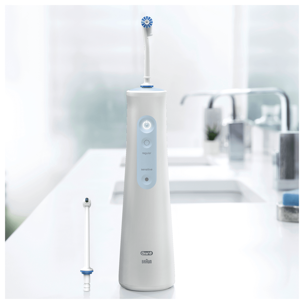 Irrigador eléctrico blanco sobre una encimera. Un cabezal de irrigador de repuesto está al lado. El fondo incluye lavabos.