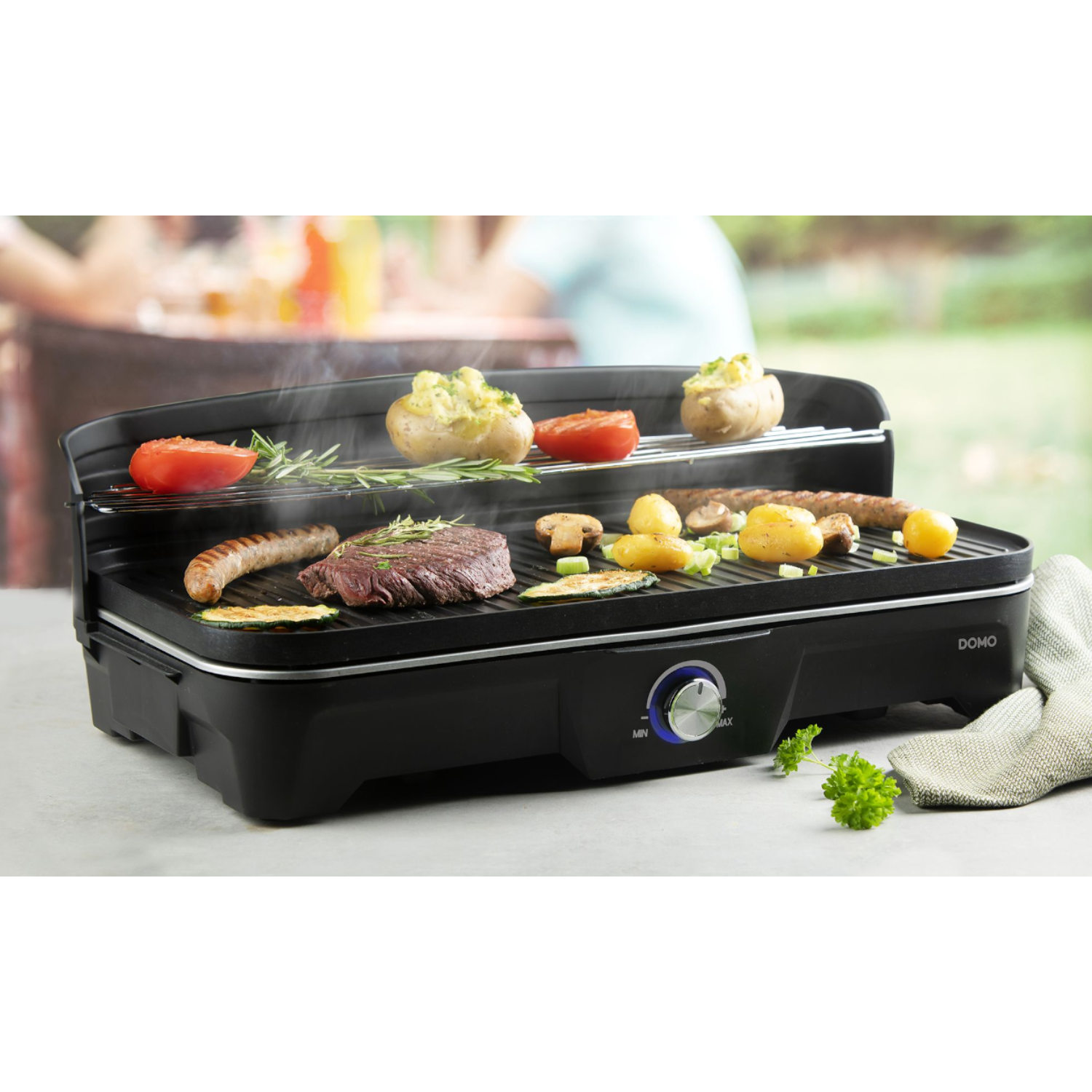 Elektrische grill met gekookt voedsel erop. Tomaten, aardappelen en worstjes zijn zichtbaar.