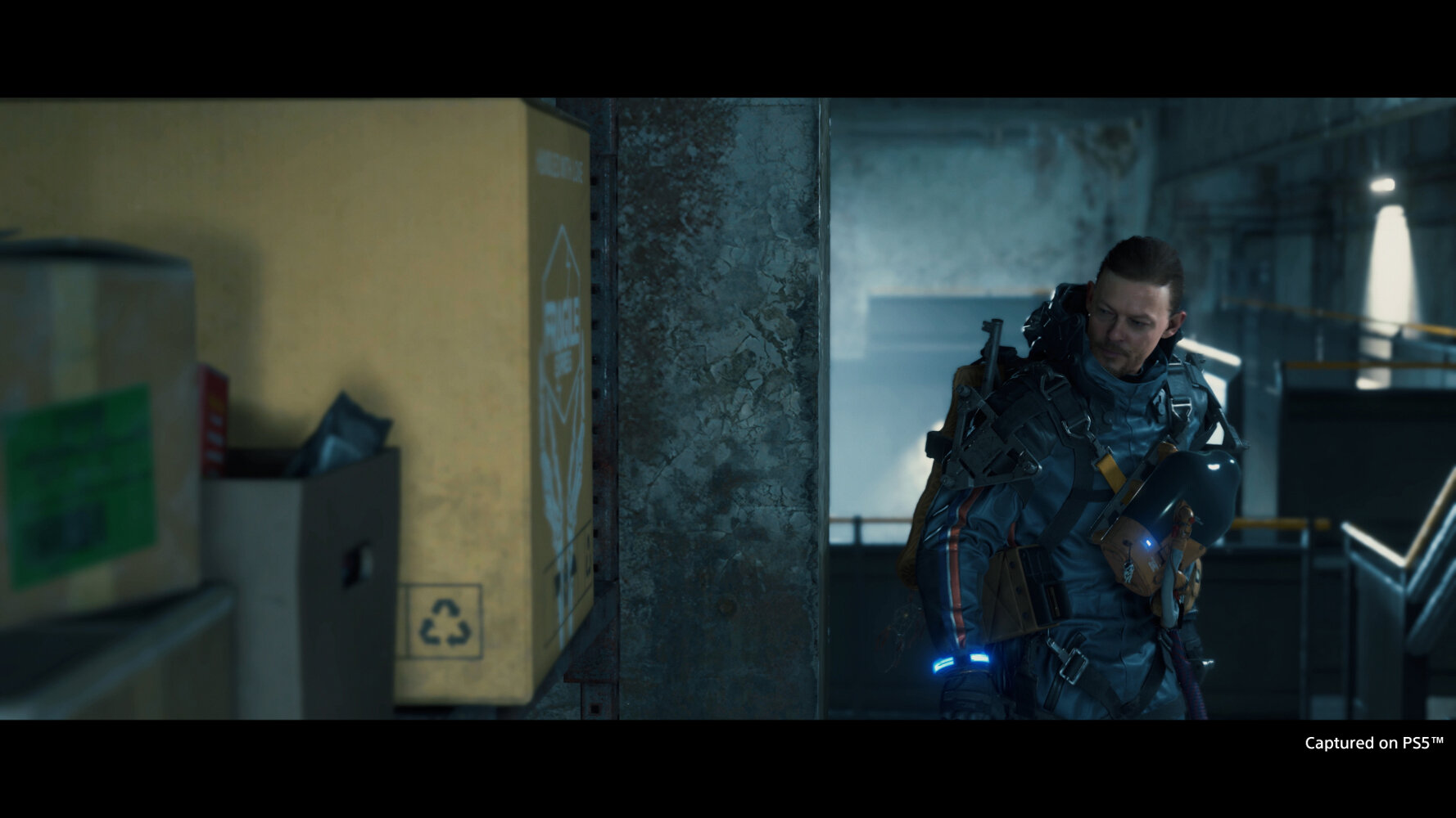 Mężczyzna w garniturze, w pobliżu ściany z pudełkami. Nosi plecak.