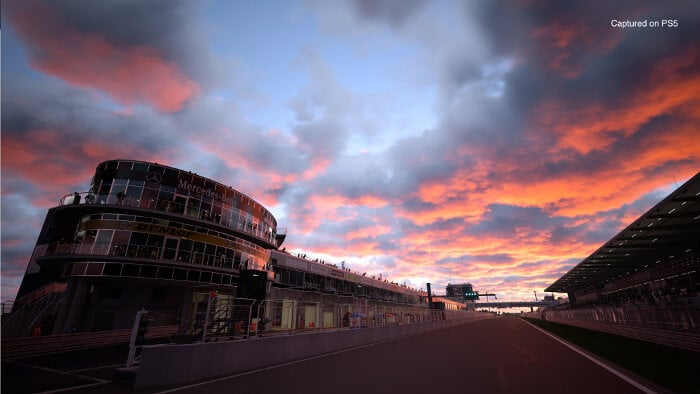 Tor wyścigowy o zachodzie słońca, z niebem z pomarańczowymi i różowymi chmurami oraz budynkiem po lewej.