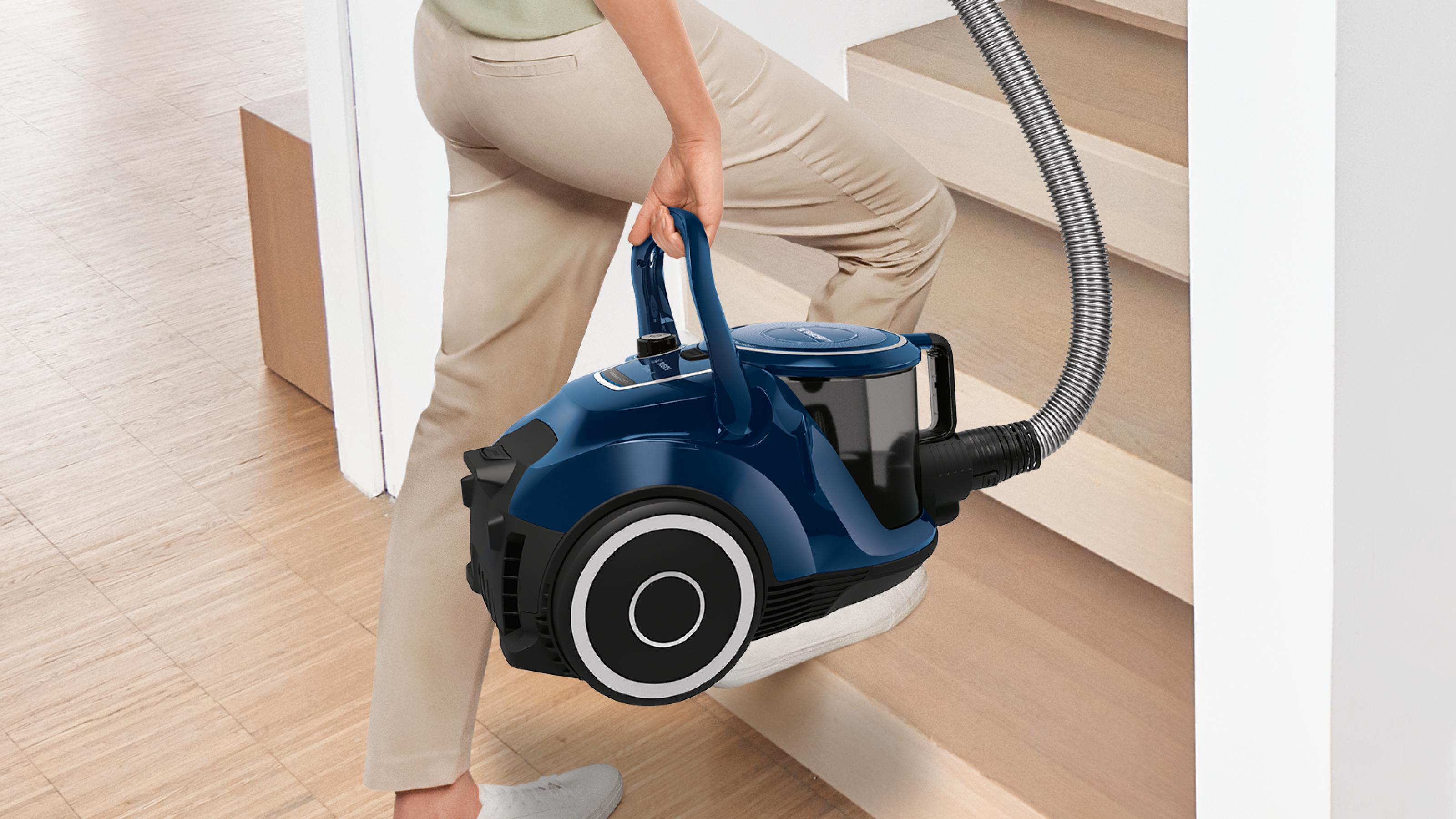 Eine Person trägt einen blauen Staubsauger eine Holztreppe hinauf, mit einer weißen Wand im Hintergrund.