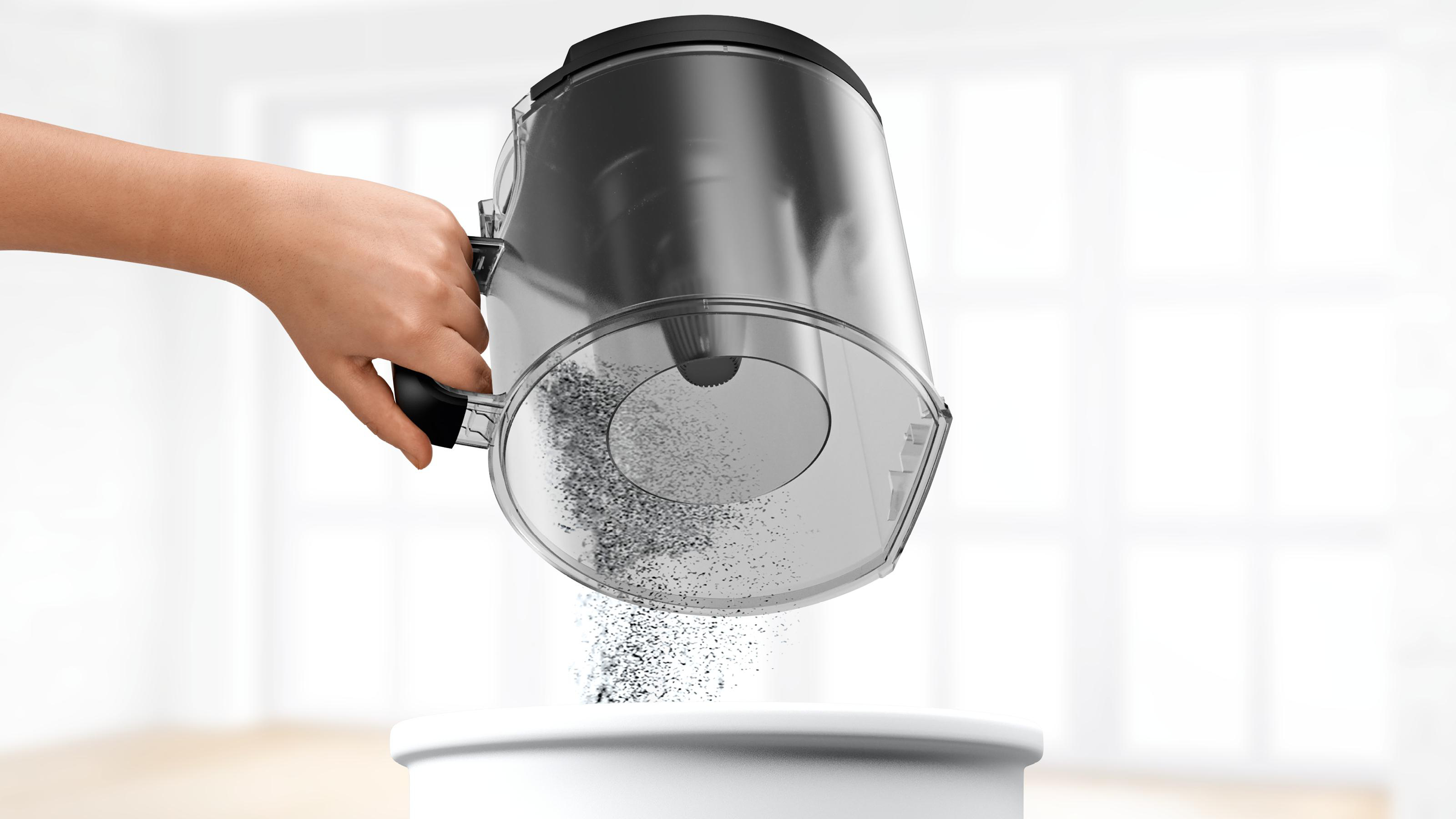 Eine Hand leert einen Staubbehälter eines Staubsaugers in einen weißen Behälter. Staubpartikel sind sichtbar.