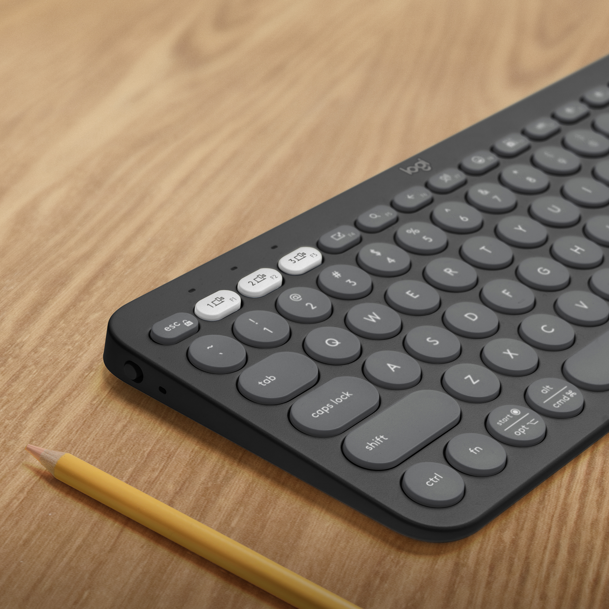 Una tastiera wireless nera e grigia con tasti rotondi e una matita gialla su una superficie di legno.