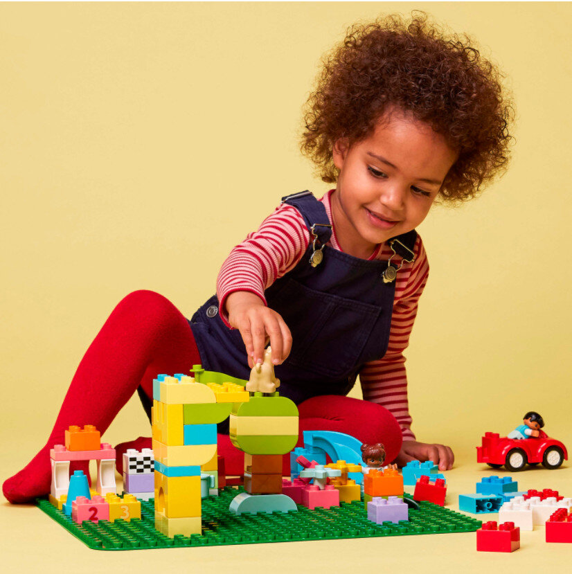 Dziecko bawiące się kolorowymi klockami i zabawkami na zielonej macie, uśmiechnięte na żółtym tle.