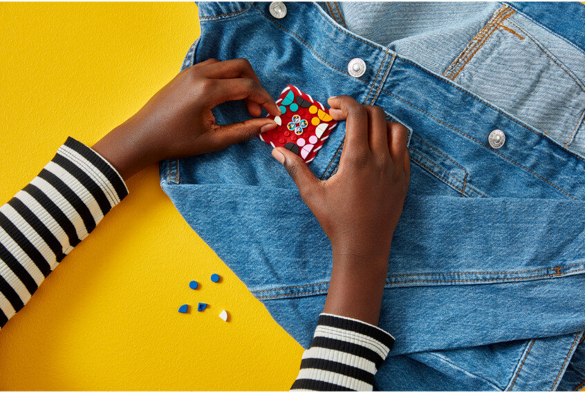 Ręce umieszczające łatkę z kwiatami Lego na dżinsowej kurtce na żółtym tle.