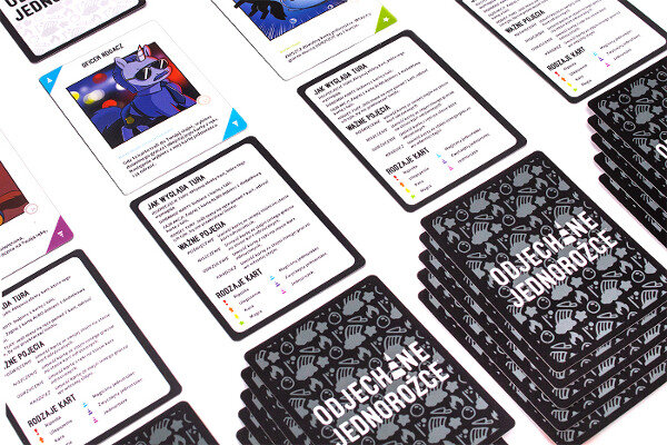 Karty do gry planszowej rozrzucone na powierzchni, z różnymi ilustracjami i tekstem w języku polskim.