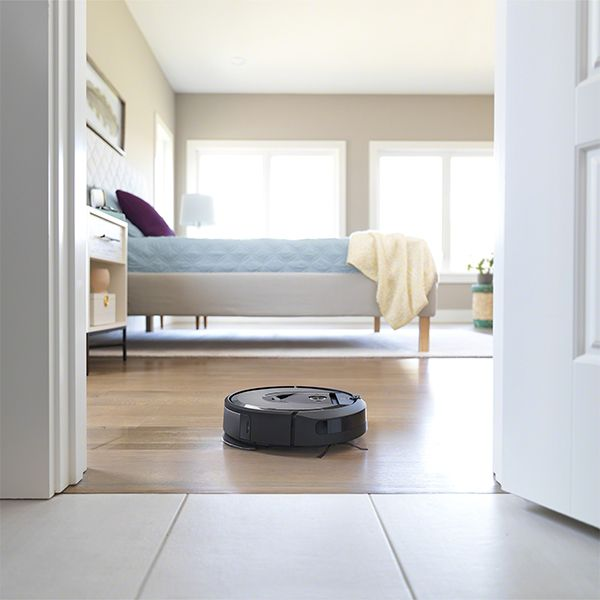 Un aspirateur robotique noir nettoie un plancher en bois dans une chambre. L'arrière-plan présente un lit.