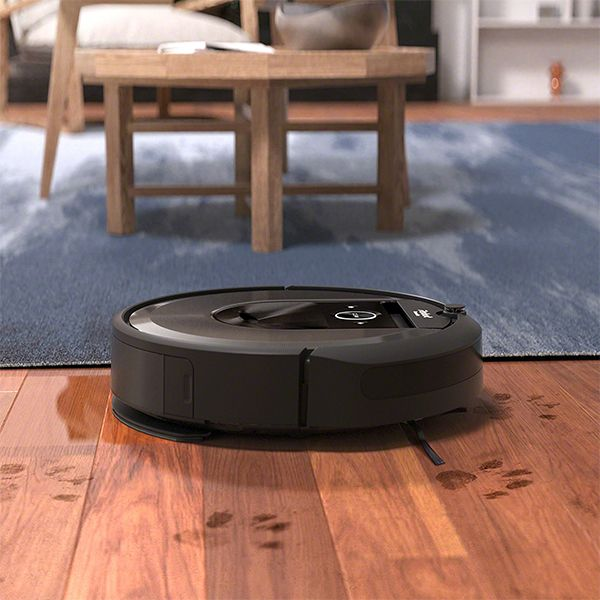 Un aspirateur robot noir sur un plancher en bois, avec des empreintes de pattes et une table en arrière-plan.