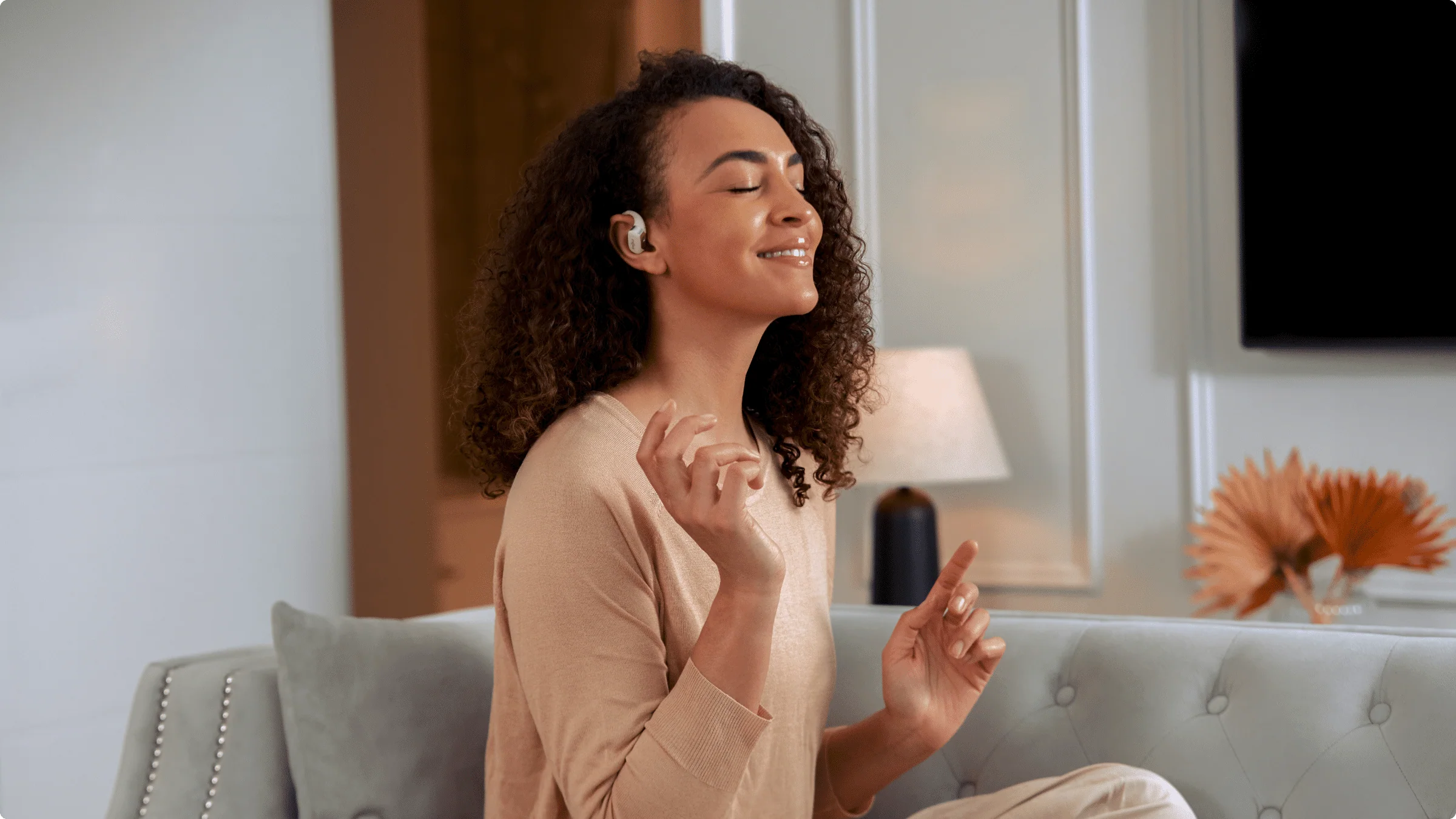 Une femme écoute de la musique avec des écouteurs sans fil, souriante et assise sur un canapé.