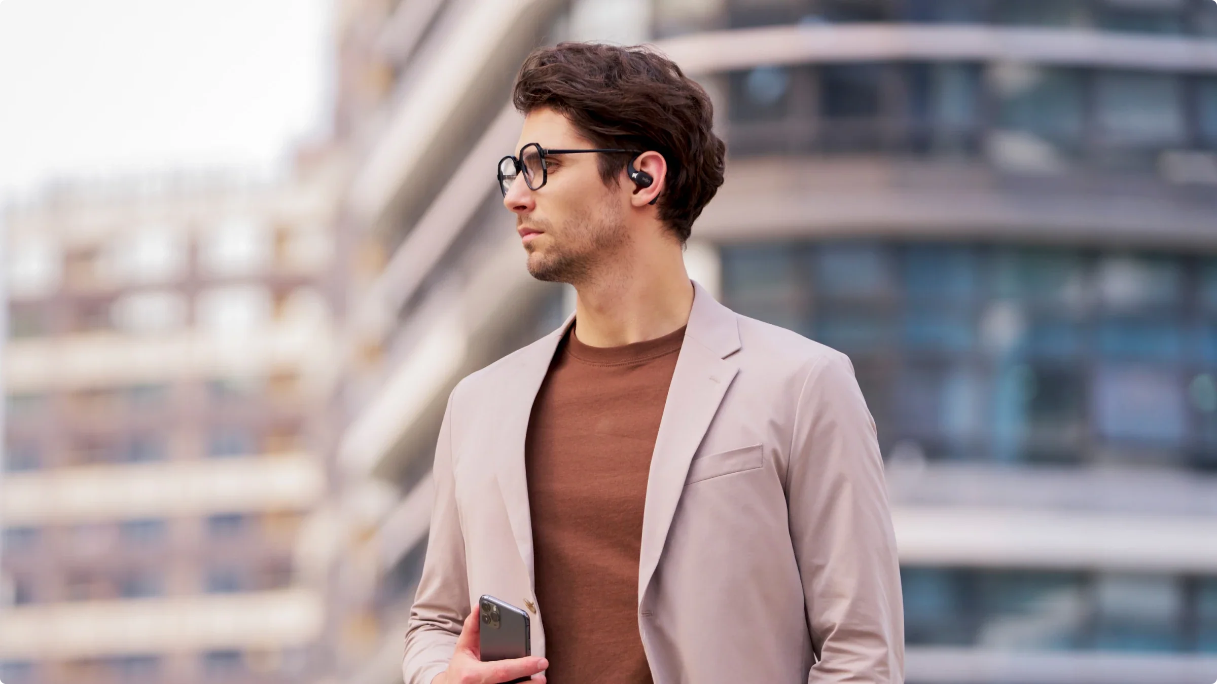 Homme avec lunettes, écouteurs, tenant un téléphone, avec fond flou.