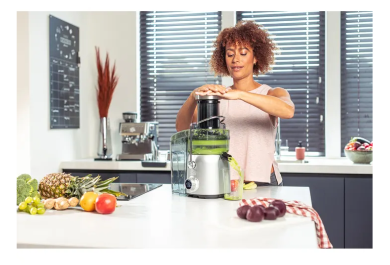 Une femme utilise une centrifugeuse sur un comptoir de cuisine avec des fruits et légumes. Une machine à café est en arrière-plan.