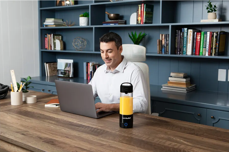 Un homme assis à un bureau utilise un ordinateur portable. Un petit mixeur est à côté d'un cahier.