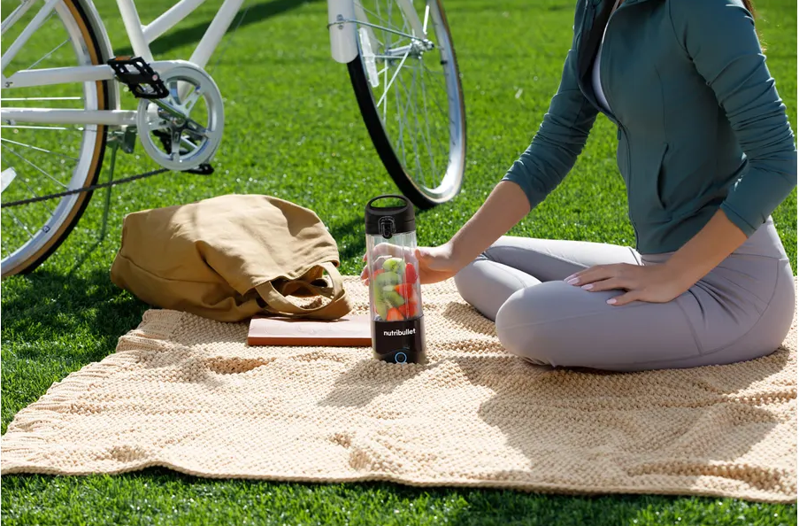 Femme sur une couverture de pique-nique avec un blender portable et des fruits, vélo derrière.