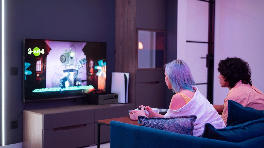 Two people on a sofa play video games on a large TV. A console and soundbar are visible.