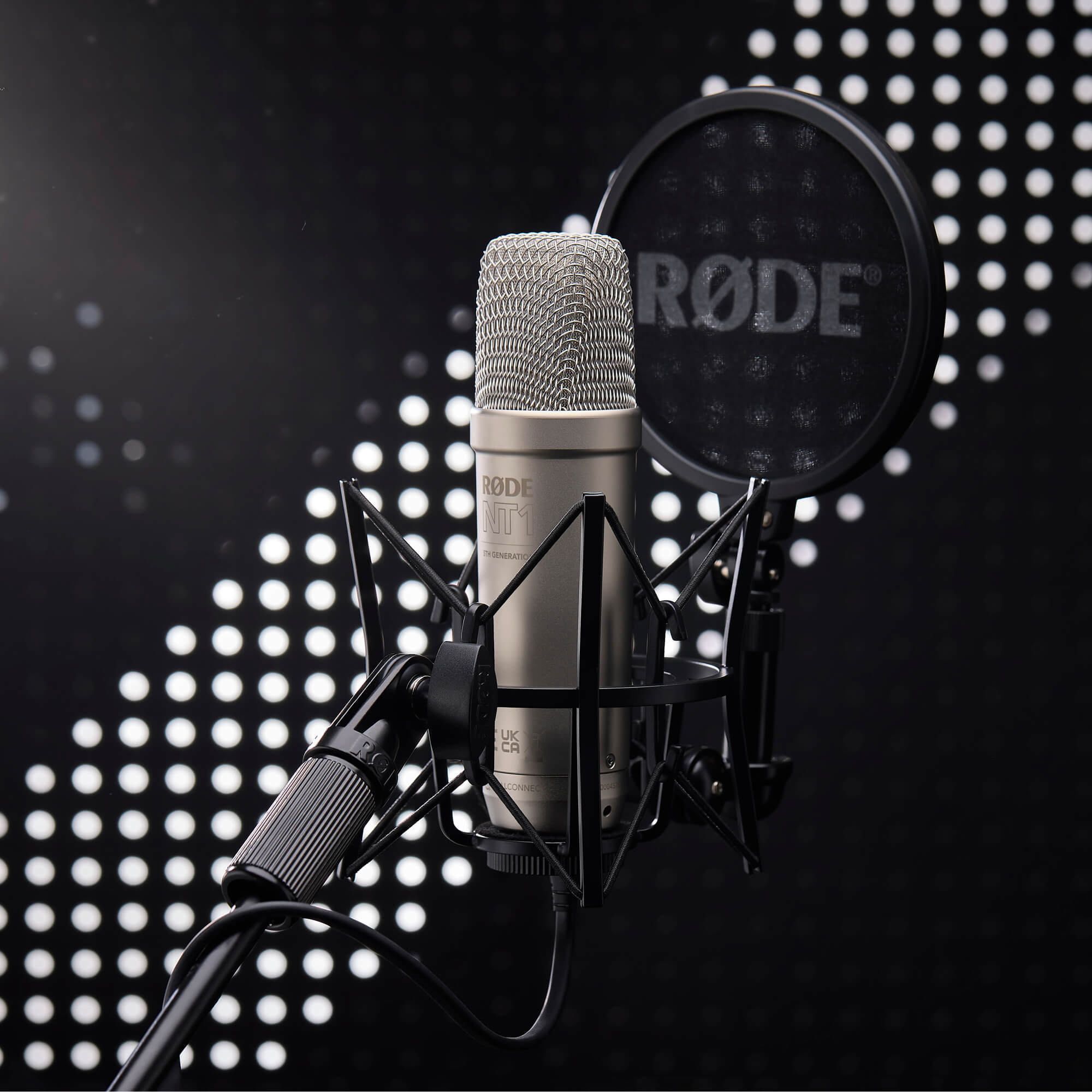 Un microphone à condensateur argenté avec filtre anti-pop noir et pied sur fond pointillé.