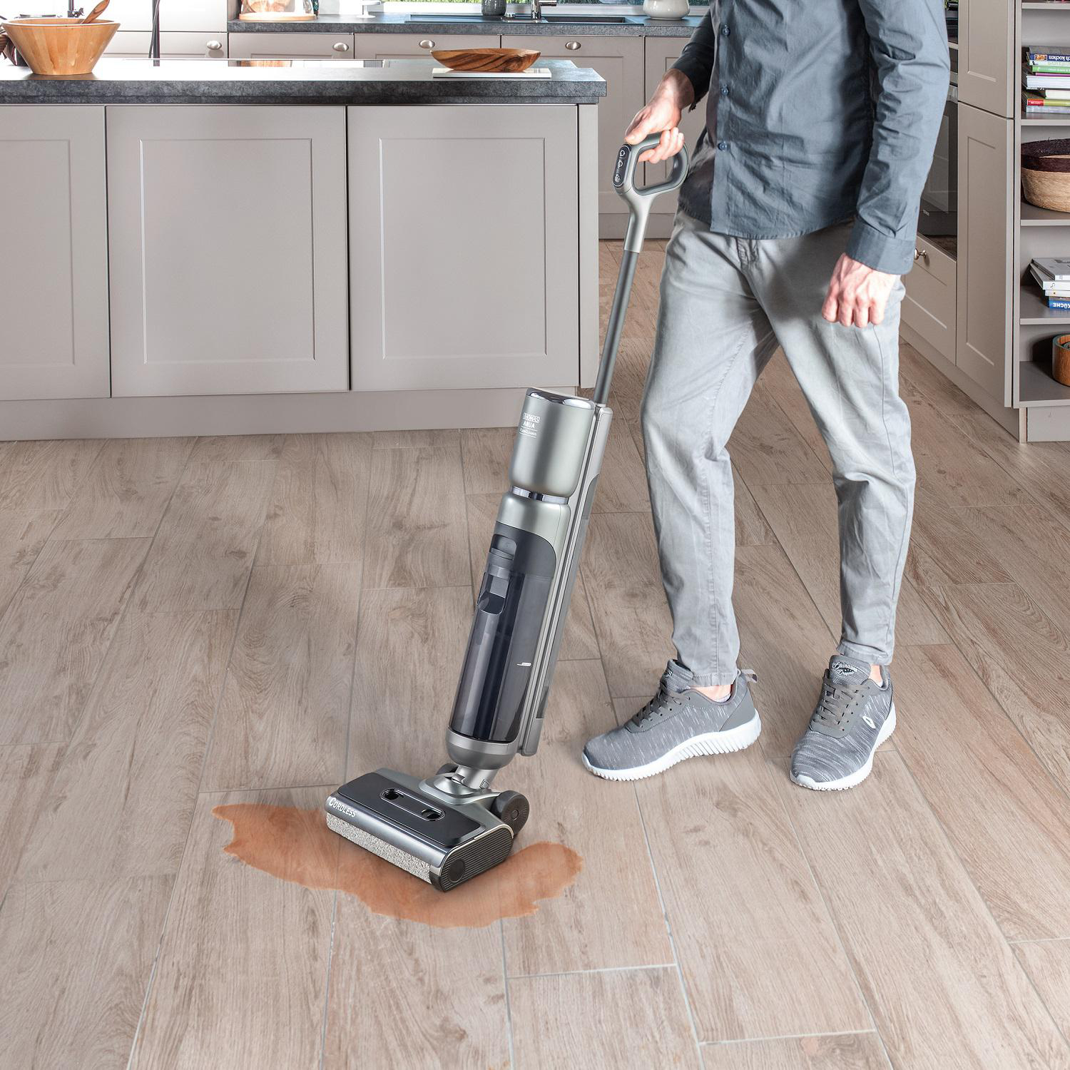 Eine Person reinigt einen Holzboden mit einem Nasssauger. Ein Wasserfleck ist sichtbar. Küchenschränke sind im Hintergrund.