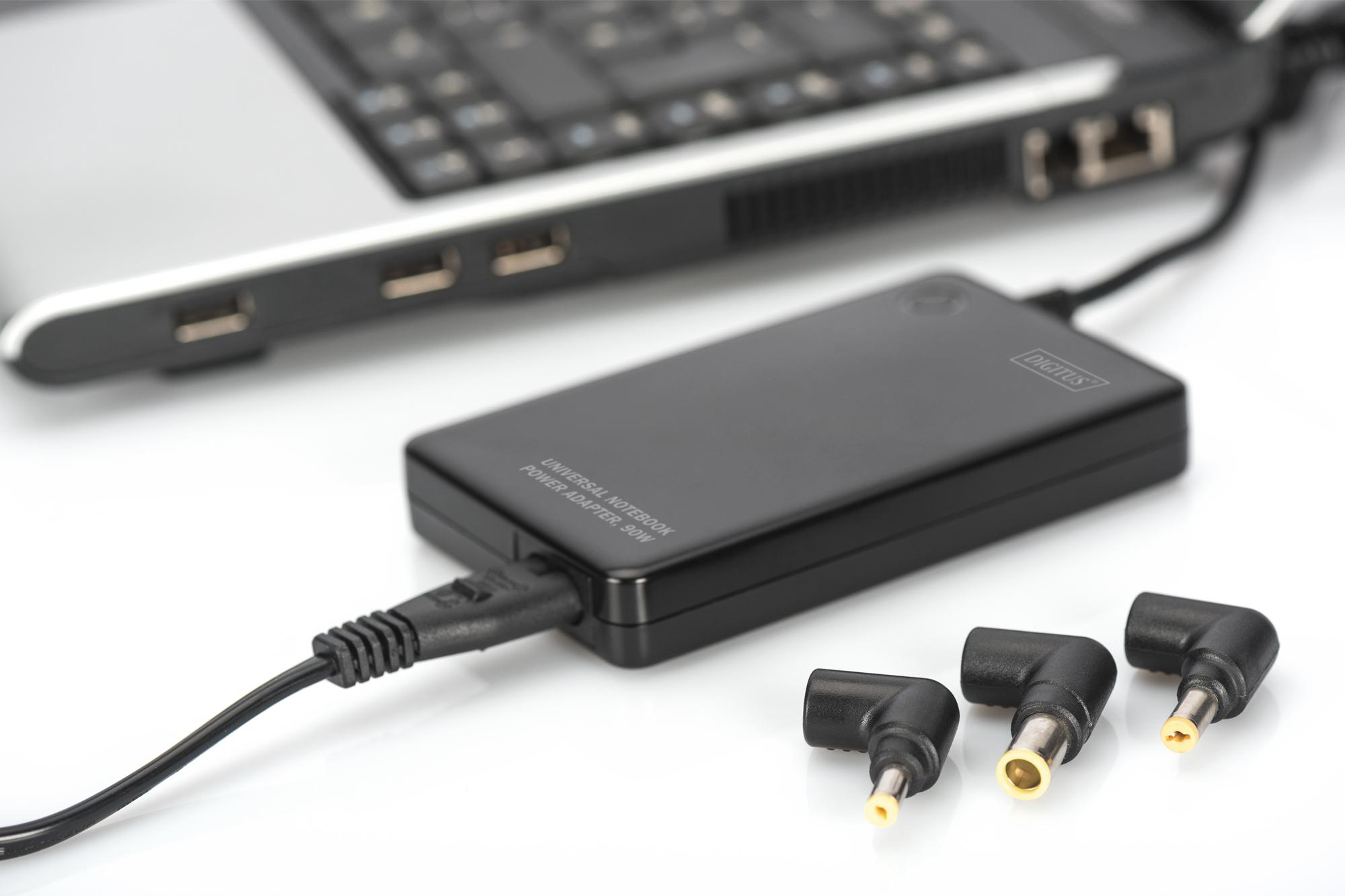 Schwarzes Notebook-Netzteil mit Anschlüssen und einem Laptop. Der Adapter ist mit dem Laptop verbunden.
