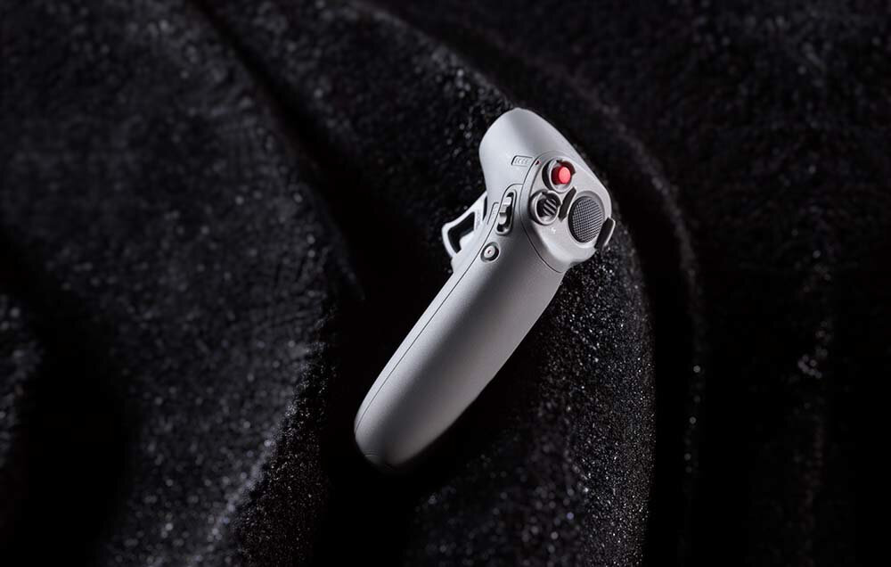 Szary kontroler ręczny z przyciskami i czerwonym światłem, leżący na ciemnej tkaninie.