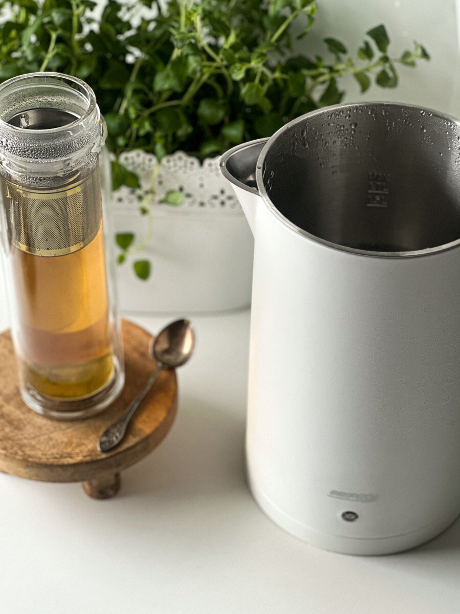 Herbata w szkle z zaparzaczem do herbaty na drewnianej podstawce obok białego czajnika.
