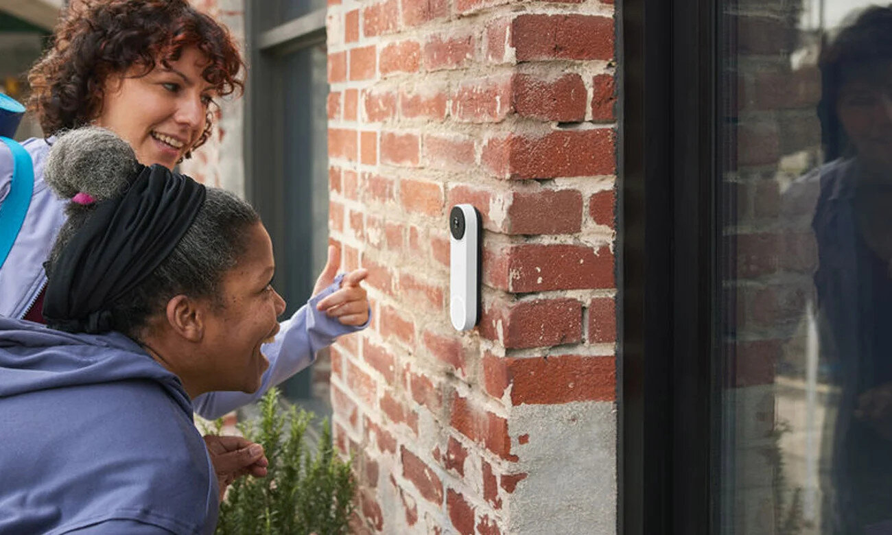 Dwie kobiety patrzą na biały dzwonek do drzwi na ceglanej ścianie. Jedna z nich wskazuje na niego.
