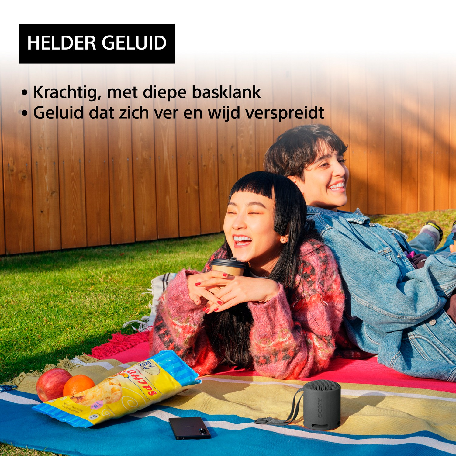 Two smiling people lying on a blanket, with snacks and a speaker in a sunny outdoor setting.