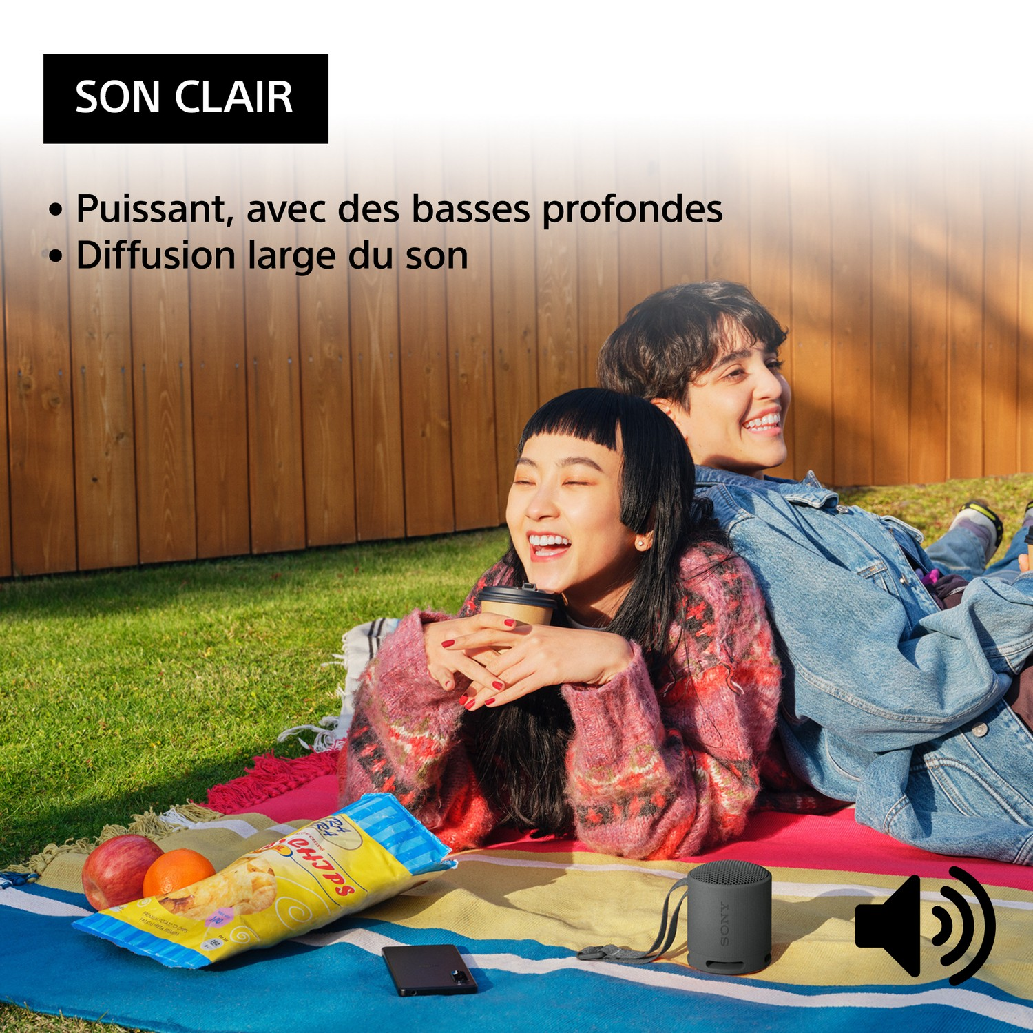 Deux personnes riant sur une couverture avec des collations, un haut-parleur et un téléphone. Clôture en bois derrière.