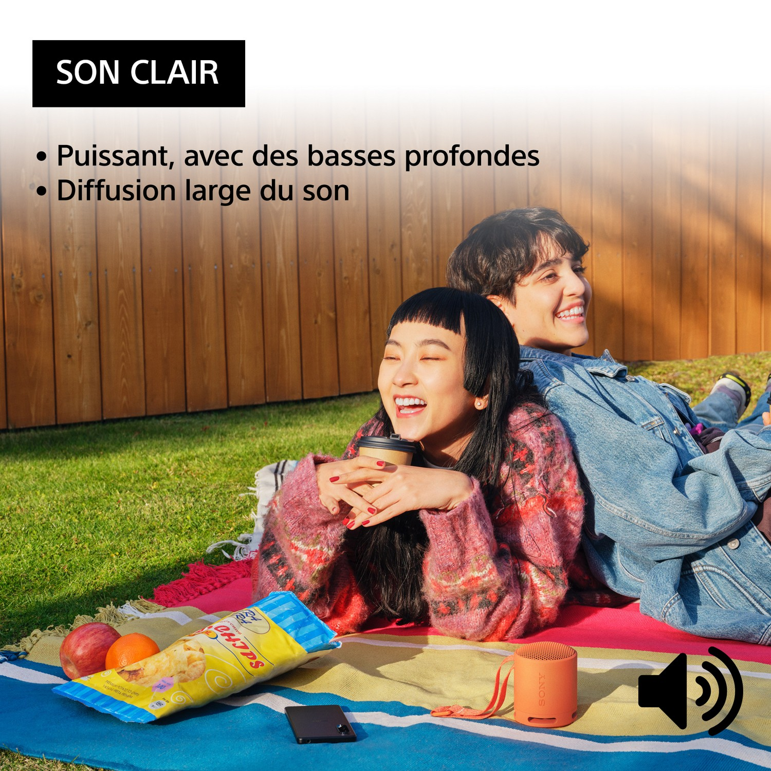 Deux personnes assises sur une couverture. Des collations et un haut-parleur sont visibles. Scène ensoleillée en extérieur.
