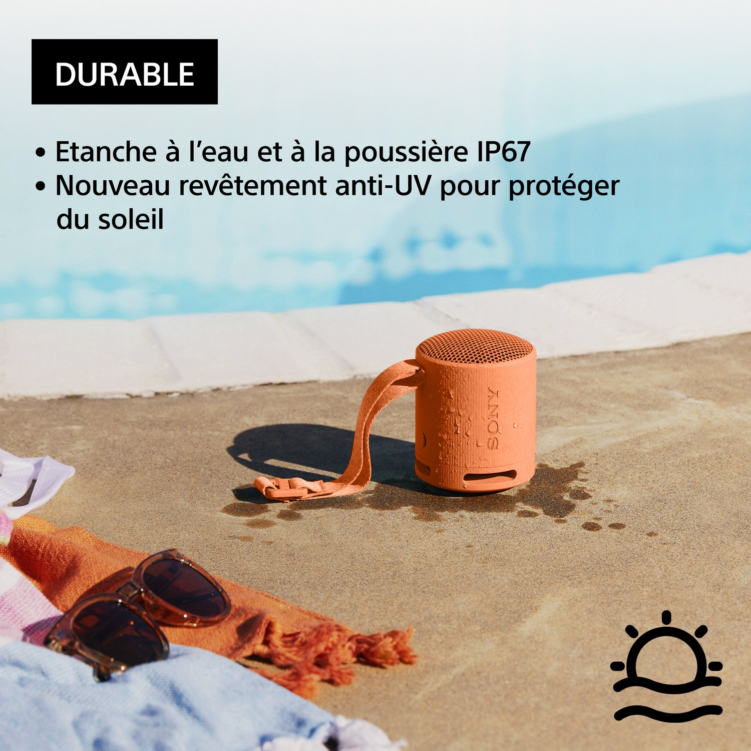 Haut-parleur portable orange à côté de lunettes de soleil et d'une serviette, avec piscine en arrière-plan.