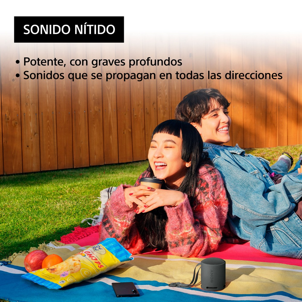 Dos personas están sentadas juntas sonriendo. Un altavoz y aperitivos son visibles en el suelo.