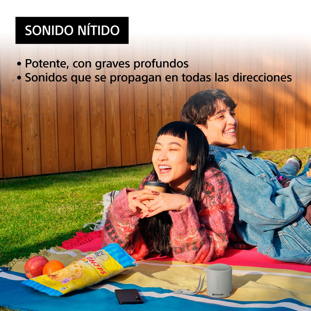 Dos personas riendo en una manta con un aperitivo y un altavoz en la hierba. Valla de madera al fondo.