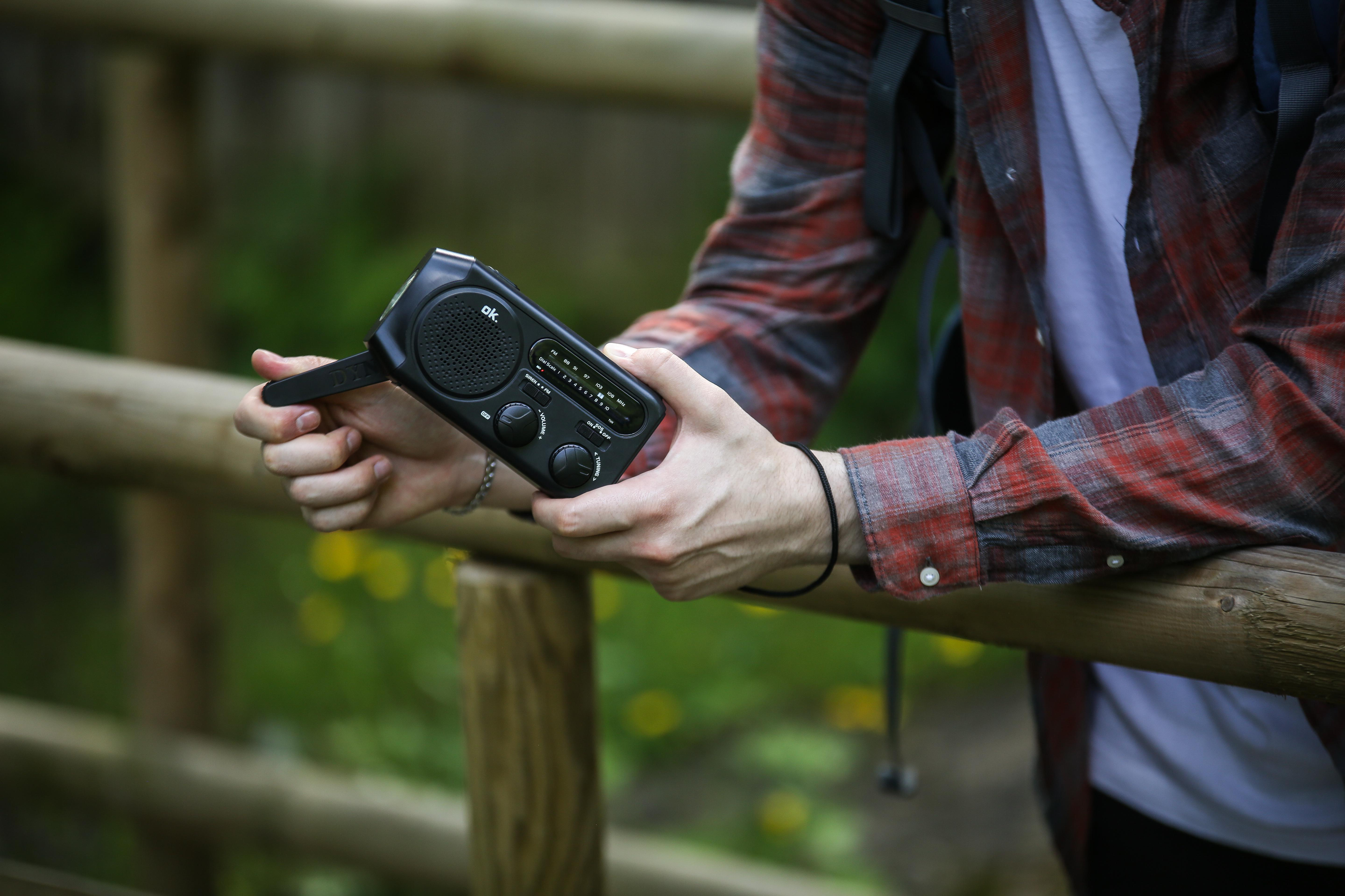 Person hält ein schwarzes Radio in der Nähe eines Holzzauns. Grüner, verschwommener Hintergrund.