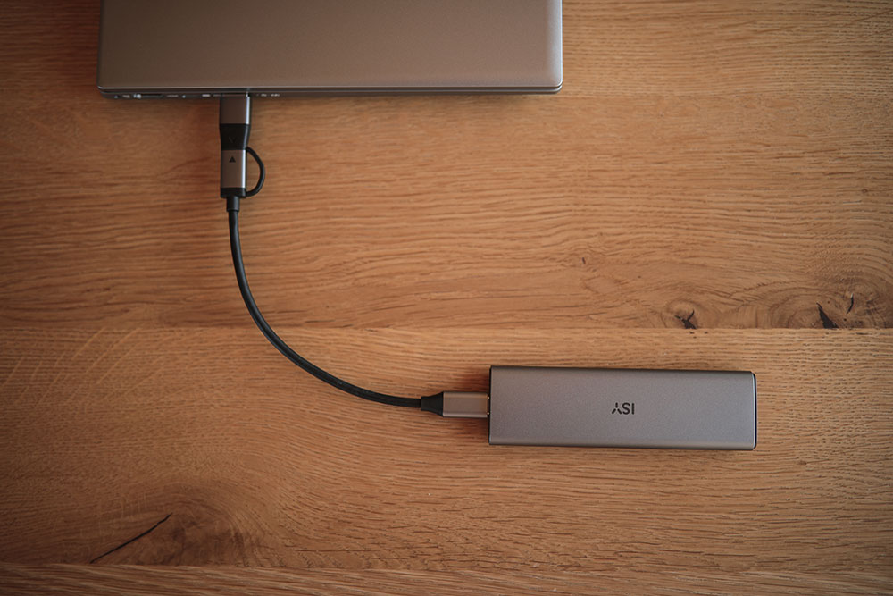 Laptop, USB-C hub, and silver external drive on wood table; black cable connecting them.