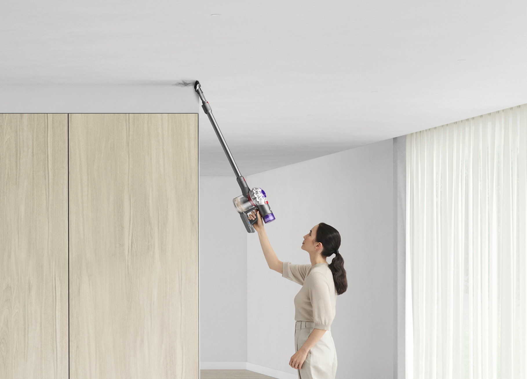 Mujer aspirando una esquina del techo con una aspiradora de mano contra una pared blanca.