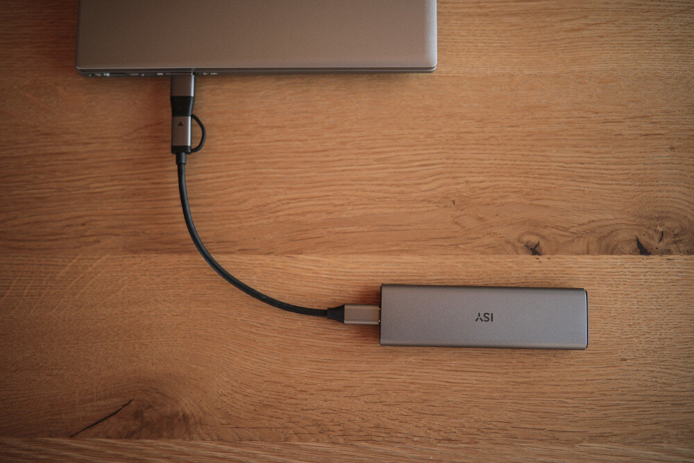 Laptop podłączony do srebrnego urządzenia ASI za pomocą kabla USB, na drewnianej powierzchni.