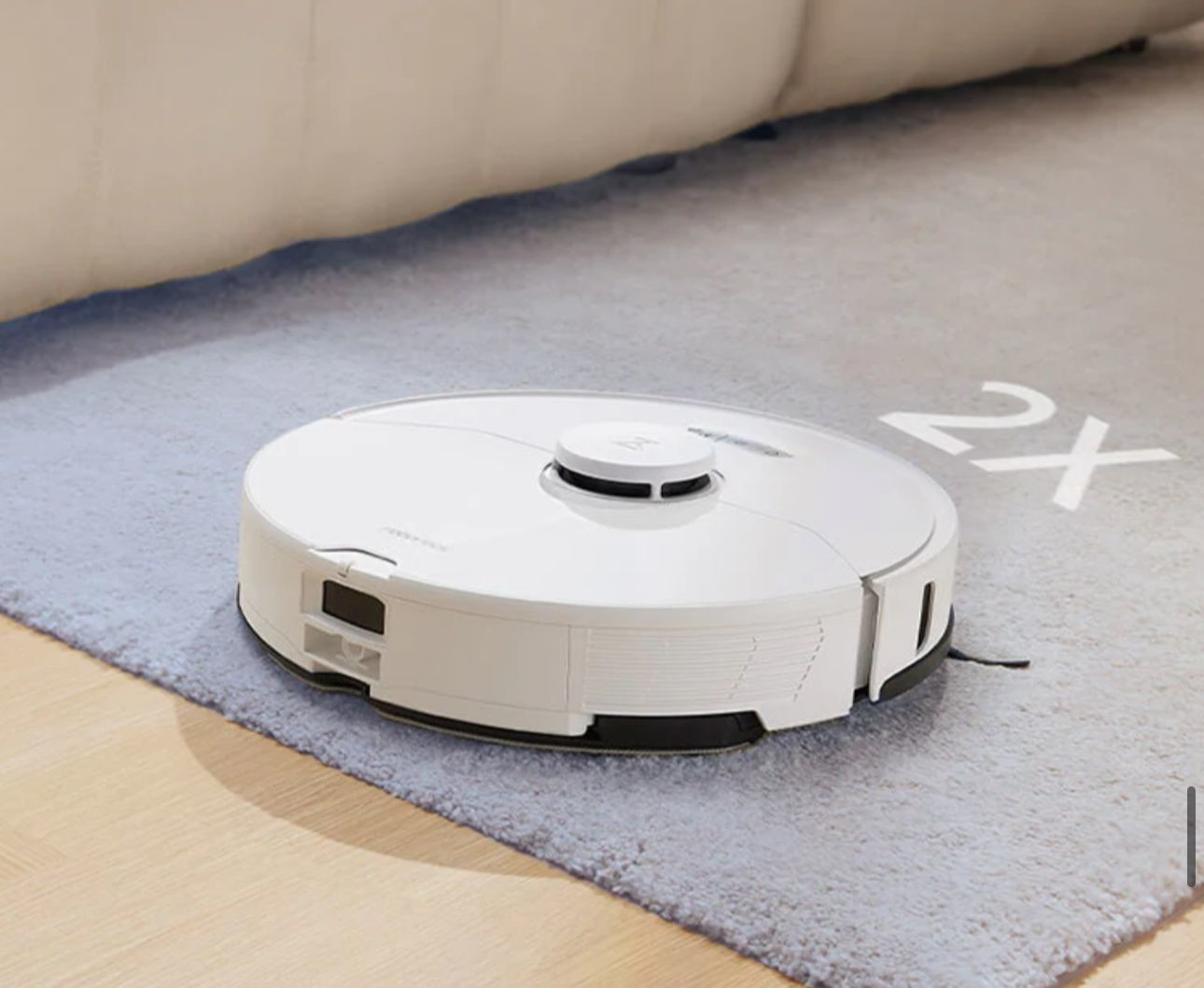 Gri bir halı üzerinde beyaz robot süpürge. 2X metni sağ tarafta.