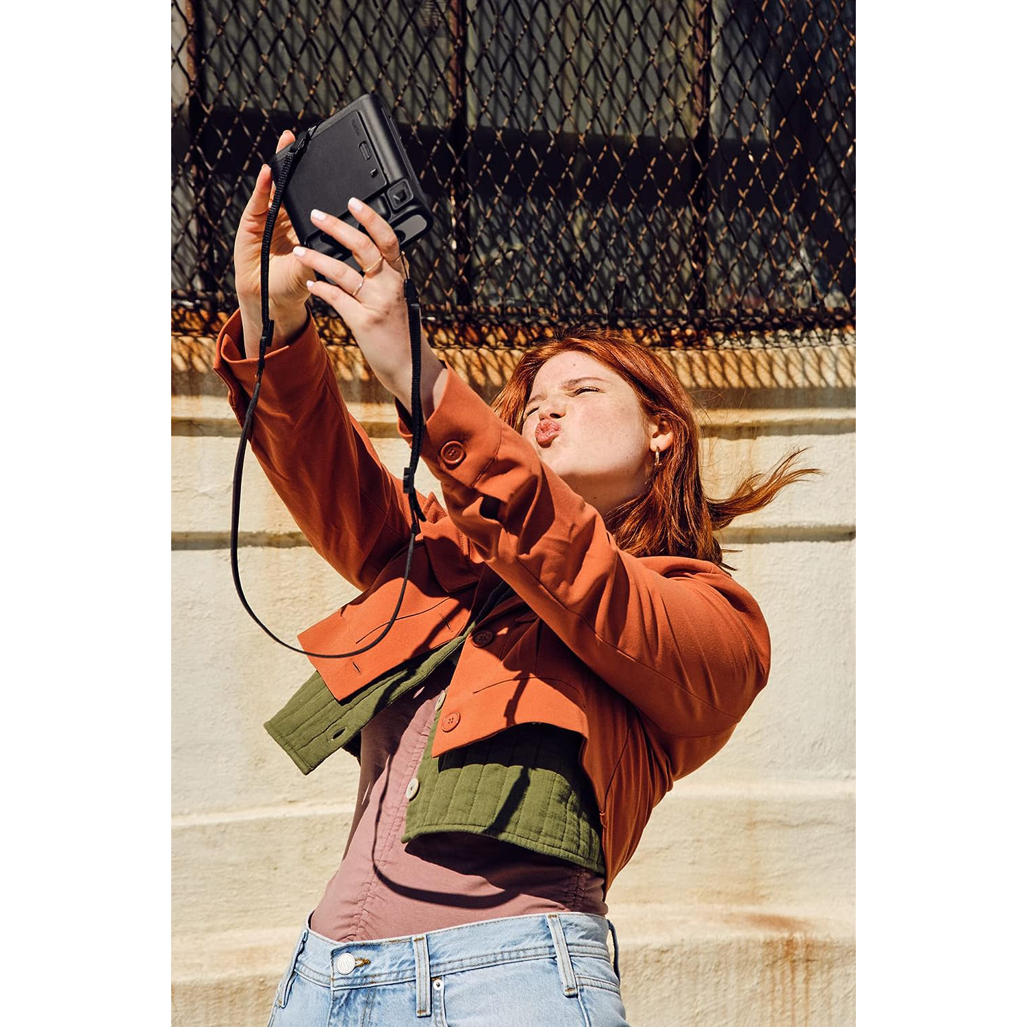Donna che si fa un selfie con una fotocamera nera, facendo il broncio. Giacca arancione, camicia verde e jeans blu.