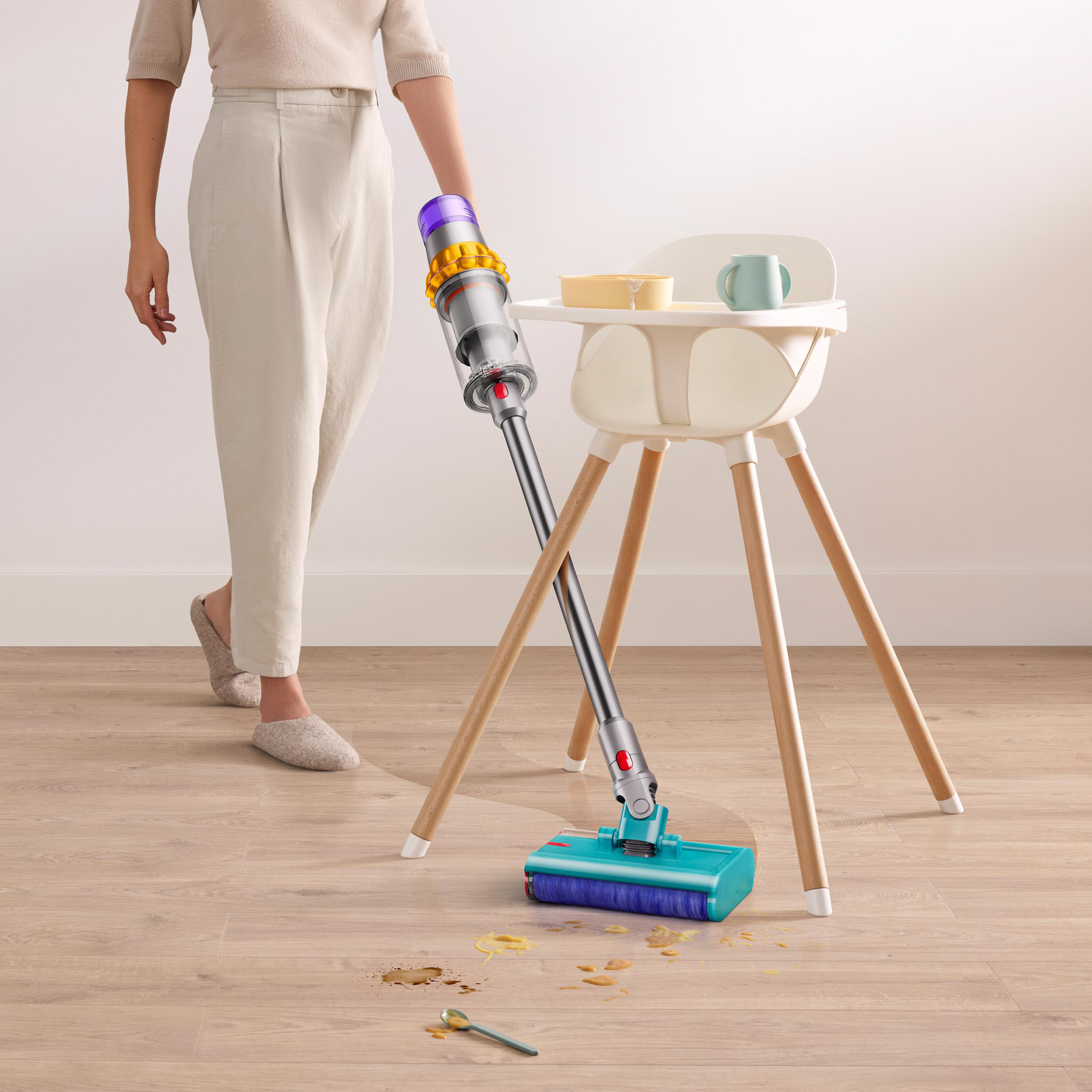 Eine Frau benutzt einen Staubsauger in der Nähe eines Babyhochstuhls, mit verschüttetem Essen auf dem Boden.