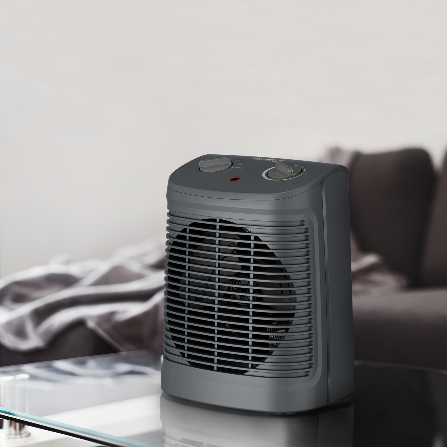 A gray electric fan heater on a glass table, with a blurred sofa and wall in the background.