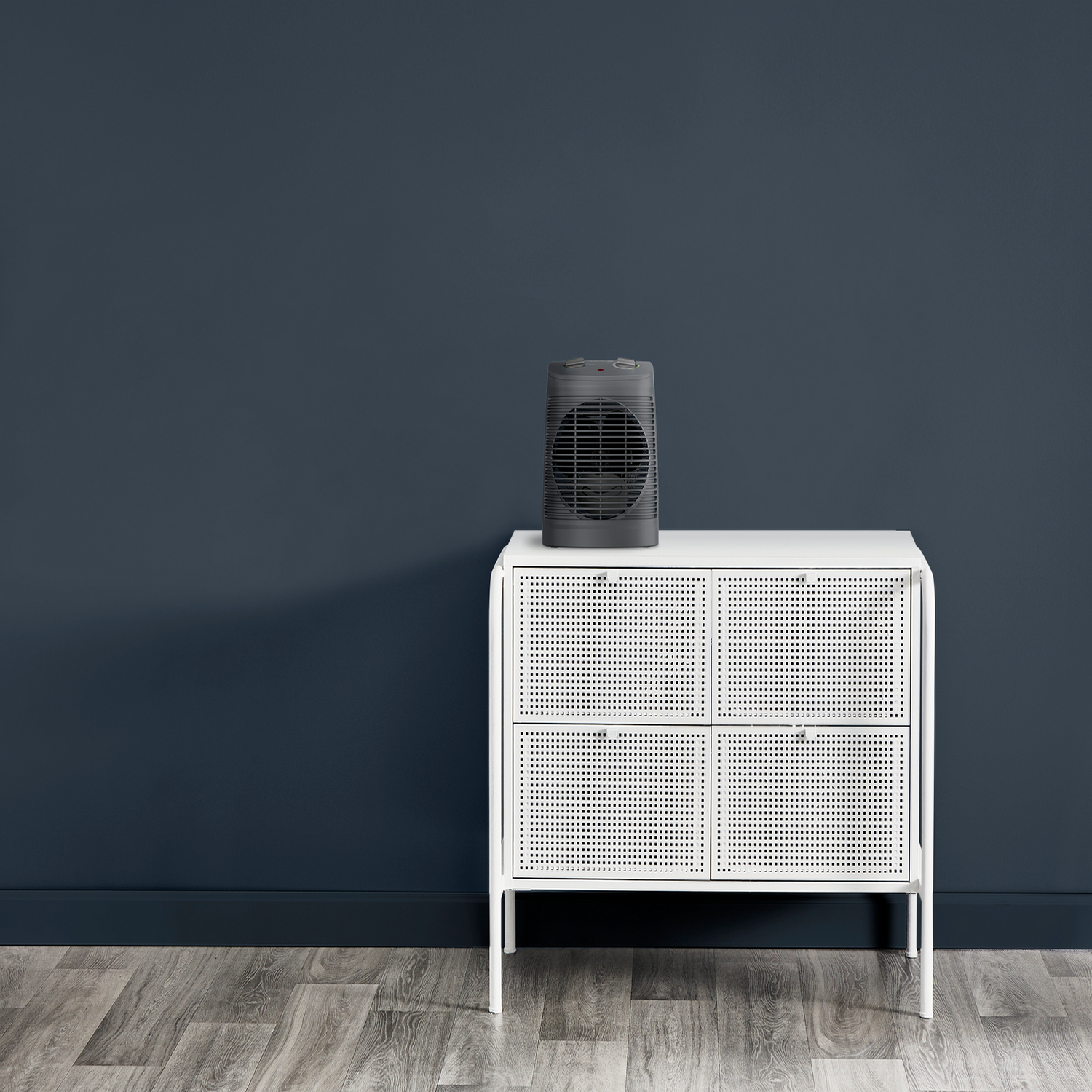 A gray fan sits on a white cabinet against a dark blue wall and wooden floor.