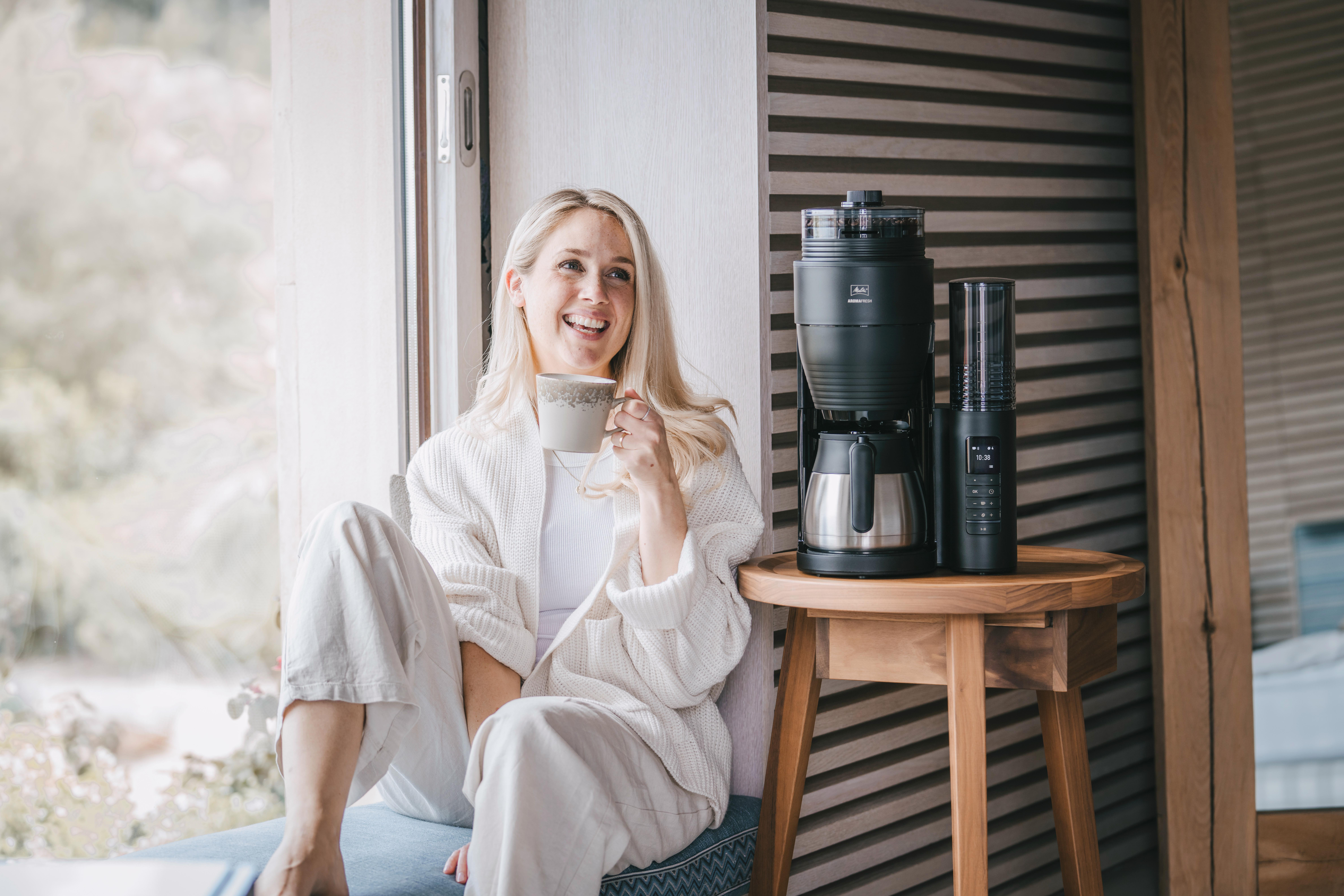 Lächelnde Frau, hält eine Tasse, sitzt am Fenster mit Kaffeemaschine.
