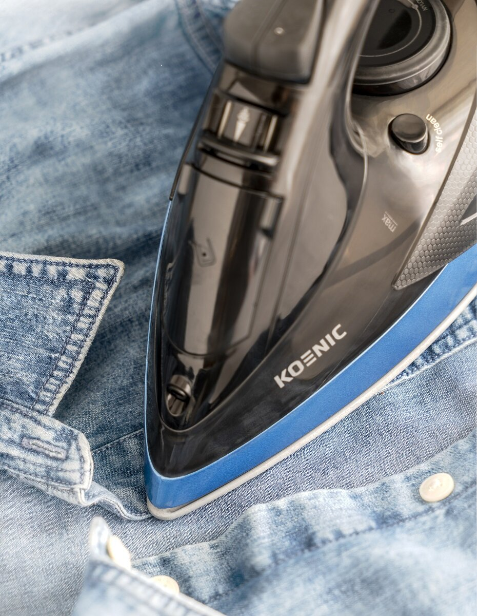 Czarno-niebieskie żelazko prasujące niebieską koszulę dżinsową, zbliżenie, pokazujące szczegóły żelazka i koszuli.