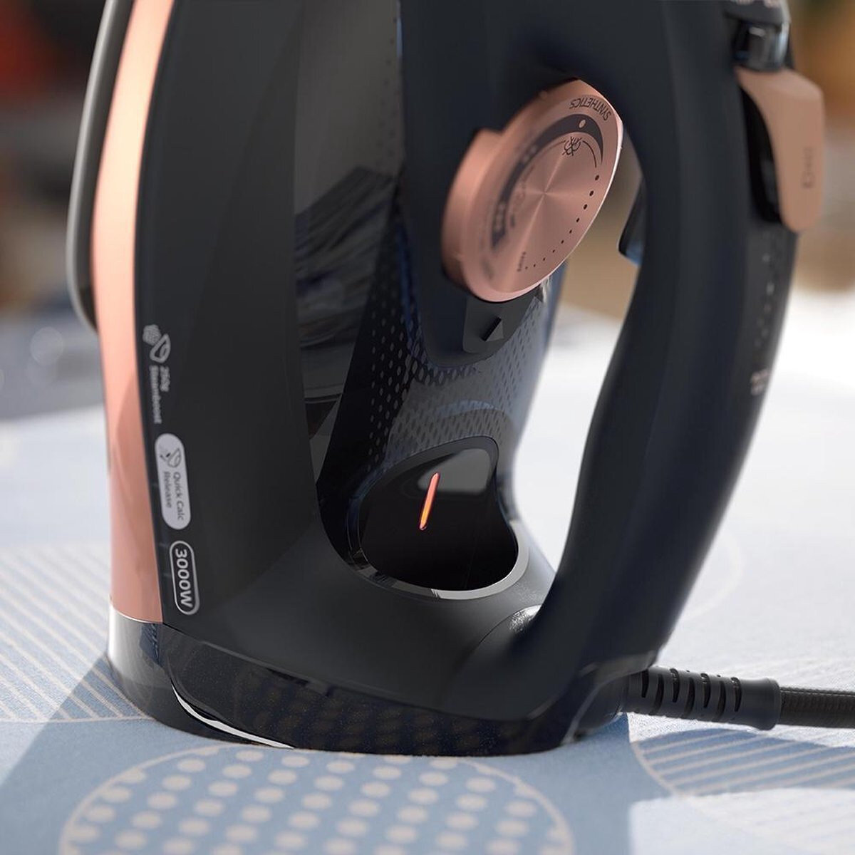 Zbliżenie czarnego i różowo-złotego żelazka z przewodem zasilającym, na niebieskiej wzorzystej powierzchni.