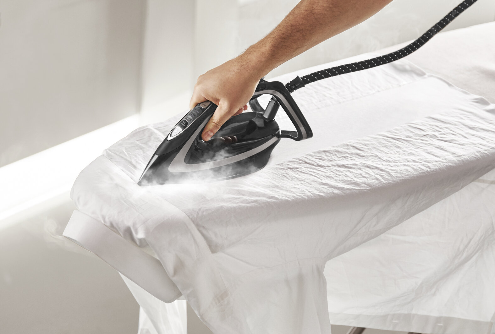 Czarne żelazko z parą prasujące białą koszulę na desce do prasowania. Widoczna dłoń.
